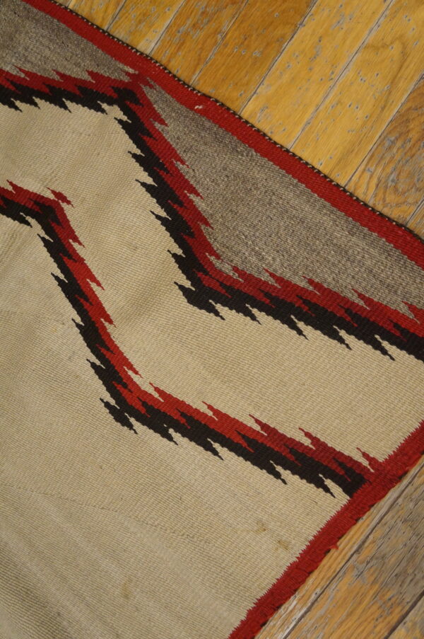 Beige flatweave rug with serrated red and black geometric designs on rustic wooden floors.