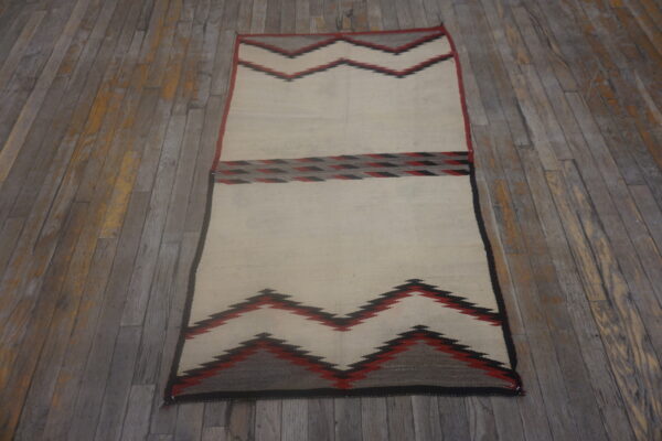 Off-white flatweave rug featuring red and black geometric zigzag bands on distressed gray wood flooring.