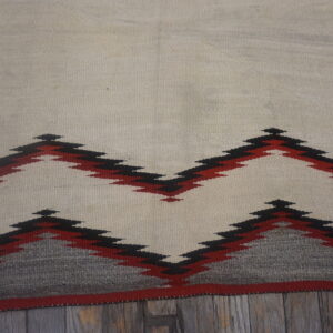 Flatweave rug with cream field and red and black geometric peaks lies on distressed wooden planks.