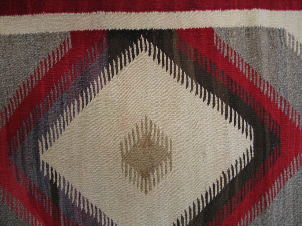 A flatwoven rug with a large central beige diamond, framed by serrated geometric shapes in red, gray, and dark brown.