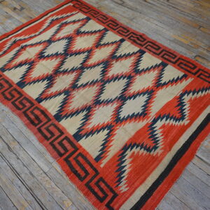 Geometric red, cream, and navy flatweave rug with a key border lies on rustic plank flooring.