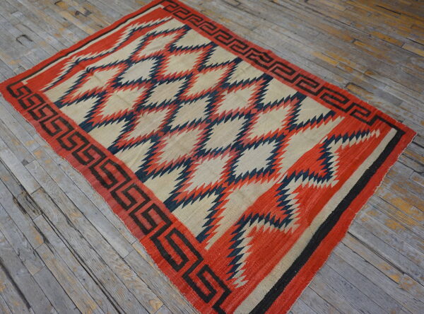 Geometric red, cream, and navy flatweave rug with a key border lies on rustic plank flooring.