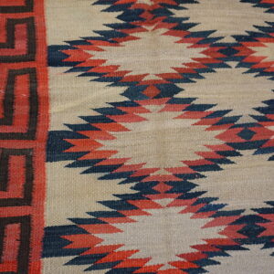 Beige flatweave rug with red and navy serrated diamonds and a red key pattern border on wood planks.