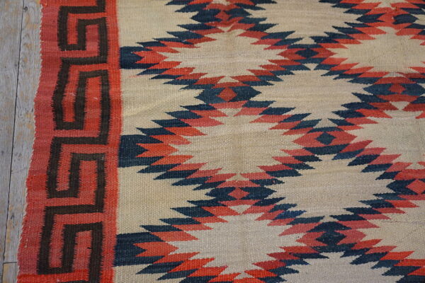 Beige flatweave rug with red and navy serrated diamonds and a red key pattern border on wood planks.