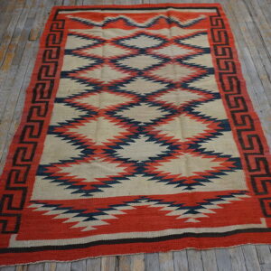 Red, navy, and tan flatweave rug with serrated diamond patterns rests on a distressed wood floor.
