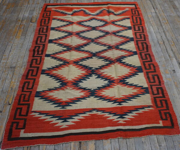 Red, navy, and tan flatweave rug with serrated diamond patterns rests on a distressed wood floor.
