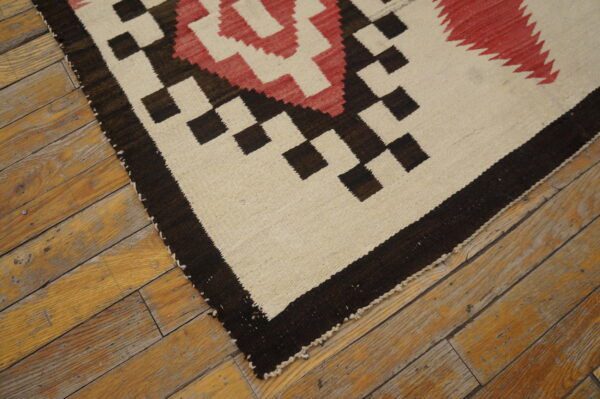 Tight-woven rug featuring geometric patterns in cream, dark brown, and red on aged wooden floorboards.