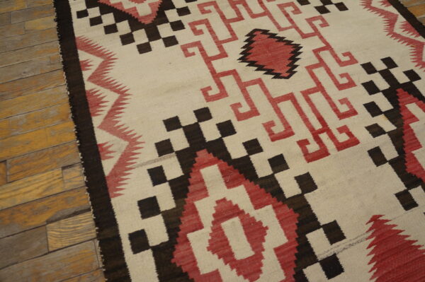 Cream flatweave rug featuring stepped geometric designs in dark brown and terracotta red on wood floor.