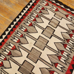 A geometric flatweave rug in cream, gray, red, and black arrow motifs lies diagonally on worn wood plank flooring.