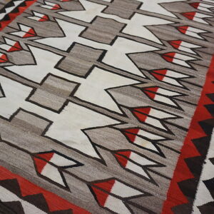 Gray flat-weave rug featuring white, red, and black geometric arrows and zigzag borders on wood flooring.