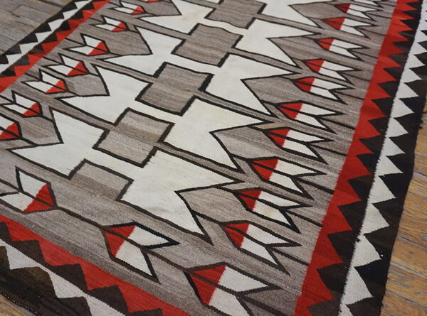 Gray flat-weave rug featuring white, red, and black geometric arrows and zigzag borders on wood flooring.
