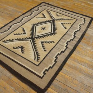 A flatwoven beige, brown, and black rug with a geometric serrated diamond pattern rests on distressed wooden floorboards.