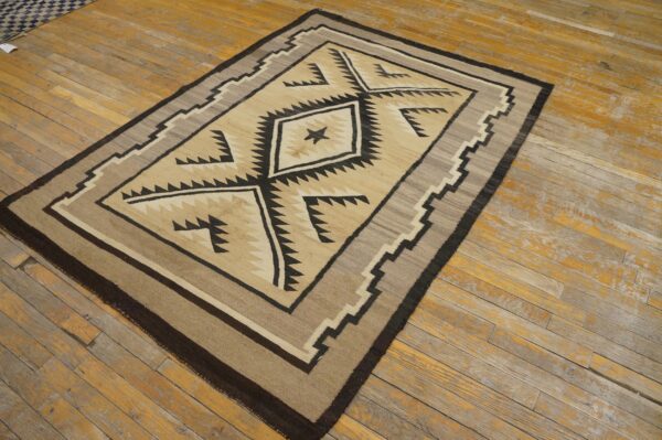 A flatwoven beige, brown, and black rug with a geometric serrated diamond pattern rests on distressed wooden floorboards.