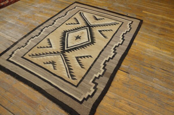 Geometric flatweave rug in tan, black, and gray with central diamond patterns rests on distressed wood flooring.