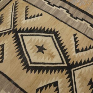 Geometric rug in tan, cream, gray, and black with serrated diamonds on worn wooden floorboards.