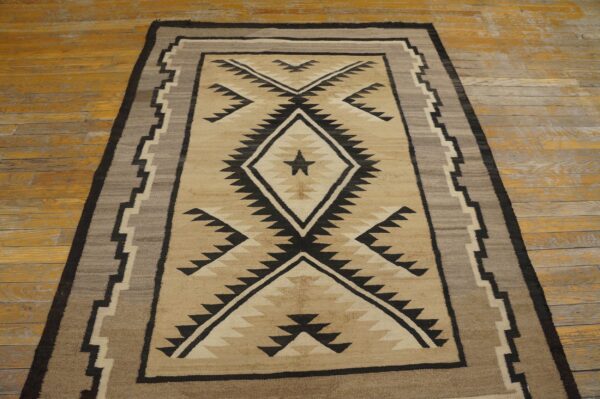 Tan and black geometric rug with a central diamond design, bordered by gray, lies on distressed wooden flooring.