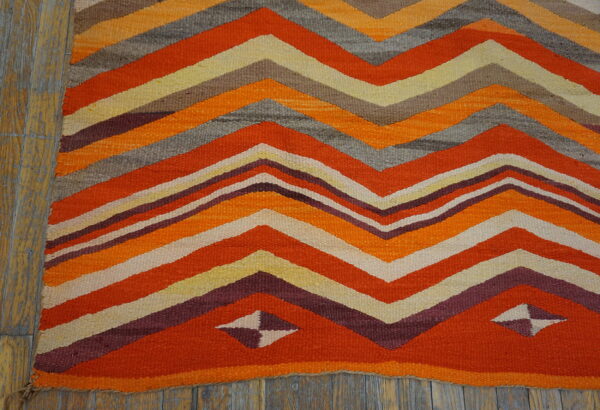 Flatweave rug with thick orange, gray, burgundy, and cream chevron stripes on weathered wood planks.