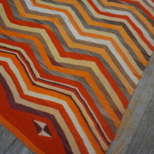A brightly colored flatweave rug featuring orange, yellow, and brown zigzag stripes rests on gray wood flooring.