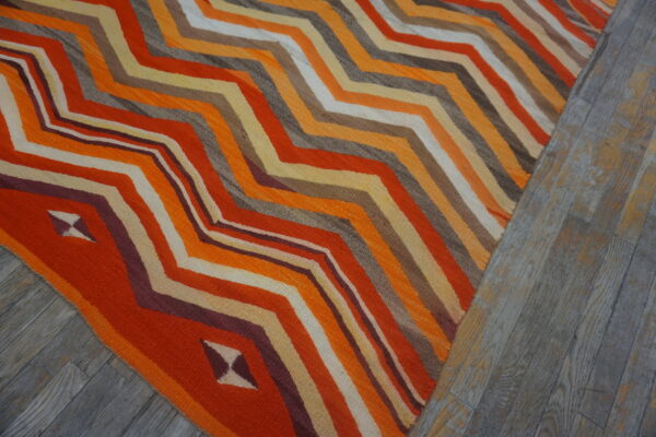 A brightly colored flatweave rug featuring orange, yellow, and brown zigzag stripes rests on gray wood flooring.