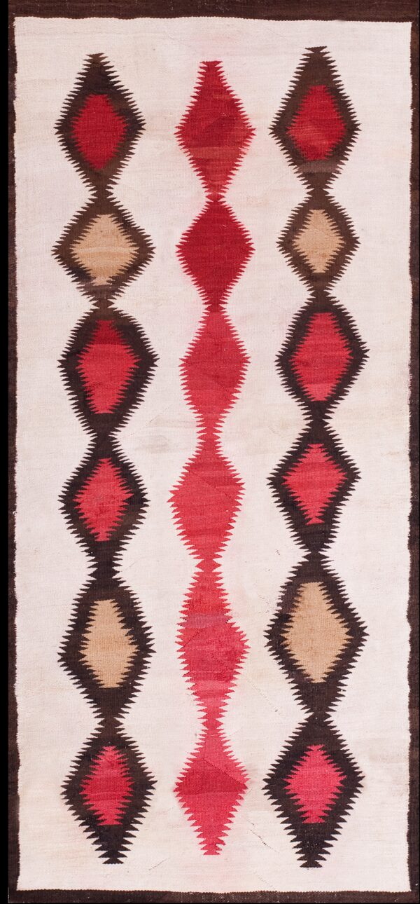 Narrow flatweave rug featuring three columns of serrated red, tan, and dark brown diamonds on a cream field over wood flooring.