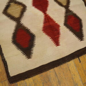 Light beige flatweave rug with vertical serrated diamonds in red, tan, and dark brown on wood floor.