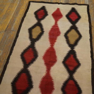 Cream flatweave rug with serrated red and tan diamond patterns outlined in dark brown on wood flooring.