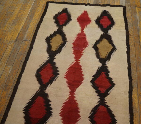 Cream flatweave rug with serrated red and tan diamond patterns outlined in dark brown on wood flooring.