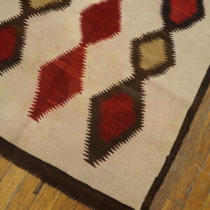 Flatweave rug featuring linked red and tan serrated diamonds on a cream field over distressed wood flooring.