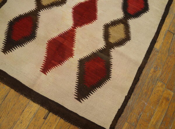 Flatweave rug featuring linked red and tan serrated diamonds on a cream field over distressed wood flooring.