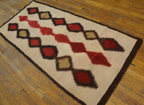 Flatwoven rug with cream ground and jagged red, brown, and tan diamonds rests on wooden plank flooring.