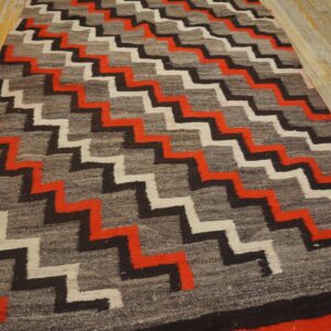 A low-pile rug with red, black, and white zigzag patterns on a gray ground rests on distressed wood flooring.