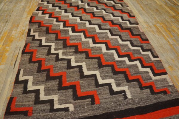 A low-pile rug with red, black, and white zigzag patterns on a gray ground rests on distressed wood flooring.