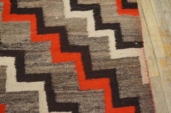 Rustic rug with bold zigzag patterns in orange, black, and cream against a mottled gray background on wood.