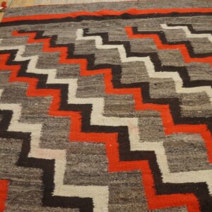Low-pile rug with interlocking orange, black, and cream zigzag geometric patterns displayed on a wood floor.
