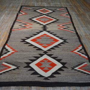 Gray rug with stacked orange, white, and black geometric diamonds lies on wide, worn wood flooring.