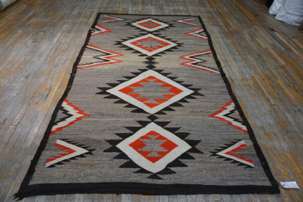 Gray rug with stacked orange, white, and black geometric diamonds lies on wide, worn wood flooring.