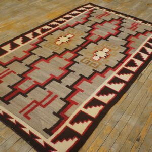 Flatweave rug with a complex gray, red, brown, and cream geometric pattern lies on worn wooden flooring.