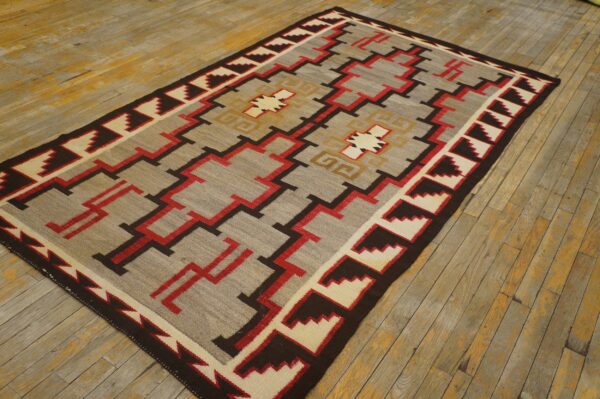 Flatweave rug with a complex gray, red, brown, and cream geometric pattern lies on worn wooden flooring.