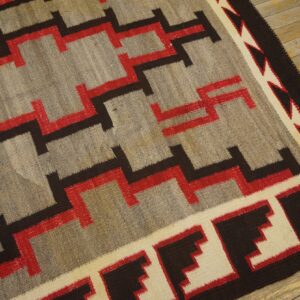 Low-pile rug in gray, red, black, and cream geometric pattern on light wooden floorboards.
