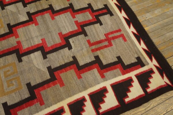 Low-pile rug in gray, red, black, and cream geometric pattern on light wooden floorboards.