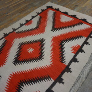 Geometric flatweave rug with a large central orange and black saw-toothed design on aged wood flooring.