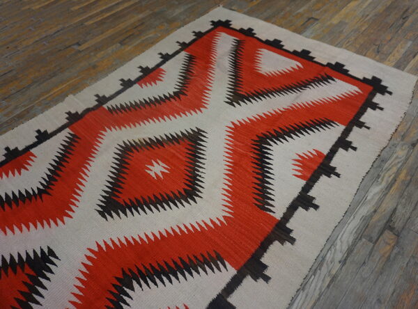 Geometric flatweave rug with a large central orange and black saw-toothed design on aged wood flooring.