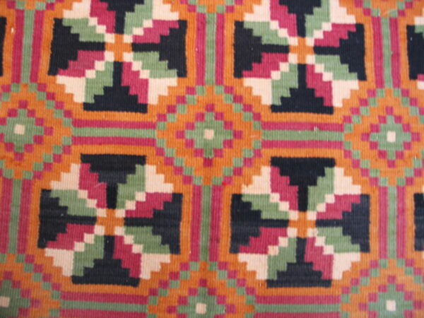 Geometric flatweave rug texture with vibrant stepped patterns in orange, magenta, black, and pale green.