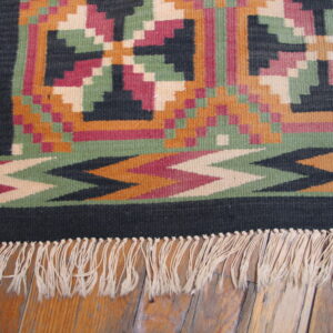A fringed geometric flatweave rug in magenta, black, green, and orange lies on a wood floor.