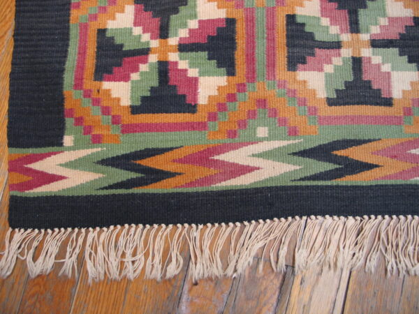 A fringed geometric flatweave rug in magenta, black, green, and orange lies on a wood floor.
