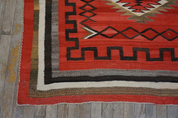 A red geometric flatweave rug with a striped border rests on distressed gray wood plank flooring.