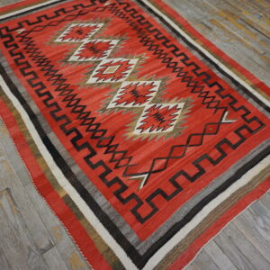 Geometric red, black, and white flatweave rug featuring diamond and fretwork patterns on worn wooden floors.