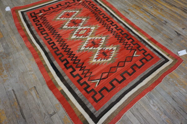 Geometric red, black, and white flatweave rug featuring diamond and fretwork patterns on worn wooden floors.