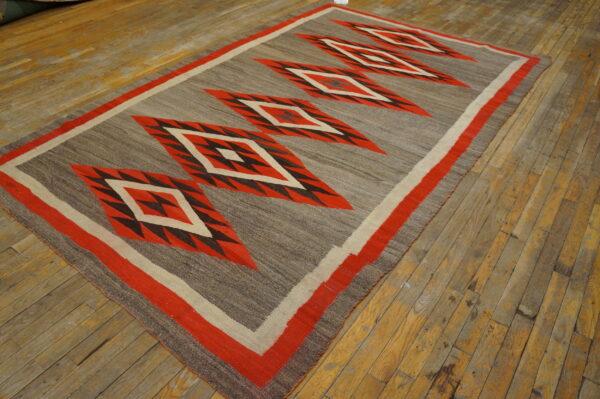 Flat-weave rug featuring large red and black geometric diamonds on a mottled gray background, resting on worn wood planks.