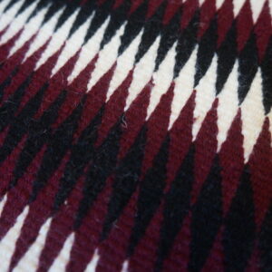 Close-up of a square rug with sharp, diagonal zigzags in cream, burgundy, and black fibers.